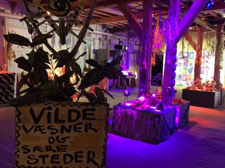 Et kig ind i udstillingen. Håndmalet skilt: Vilde Væsner og Sære Steder, ultraviolet lys, børns vilde væsner lavet af forskellige materialer på podie